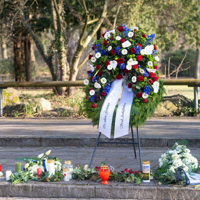 Bild vergr&ouml;&szlig;ern: Kranzniederlegung zum Gedenken an die Opfer der Gewalttat im Schöntal. Kranz ist aufgestellt, davor Blumen und Kerzen.