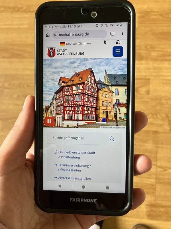 Handy in einer Hand, auf dem Handy geöffnet die Webseite der Stadt Aschaffenburg. 