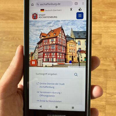 Handy in einer Hand, auf dem Handy geöffnet die Webseite der Stadt Aschaffenburg. 