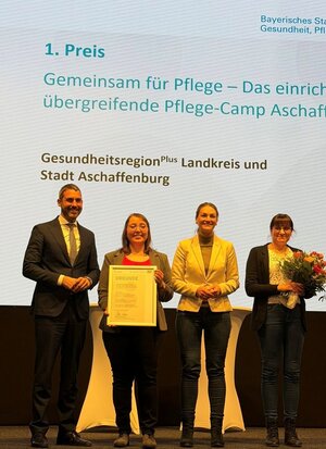 Bild vergr&ouml;&szlig;ern: Landrat Dr. Alexander Legler, Geschäftsstellenleiterin der Gesundheitsregionplus Monika Gabel, Staatsministerin Judith Gerlach und Kathleen Tobi (Lehrkraft für Berufspädagogik an der BiG und Mitorganisatorin des Pflegecamps aus dem Ausbildungsverbund Pflege Aschaffenburg) stehen auf der Bühne mit dem Preis und freuen sich.