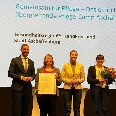 Landrat Dr. Alexander Legler, Geschäftsstellenleiterin der Gesundheitsregionplus Monika Gabel, Staatsministerin Judith Gerlach und Kathleen Tobi (Lehrkraft für Berufspädagogik an der BiG und Mitorganisatorin des Pflegecamps aus dem Ausbildungsverbund Pflege Aschaffenburg) stehen auf der Bühne mit dem Preis und freuen sich.